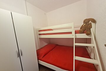 A cozy room features white bunk beds with vibrant red bedding, a matching white wardrobe, and a brown stuffed animal resting on the top bunk.