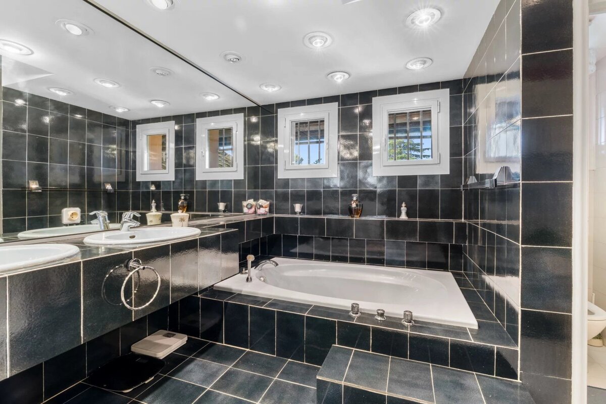 Modern bathroom featuring dark tiles, a white built-in bathtub, double sinks, a large mirror, and bright ceiling lights.