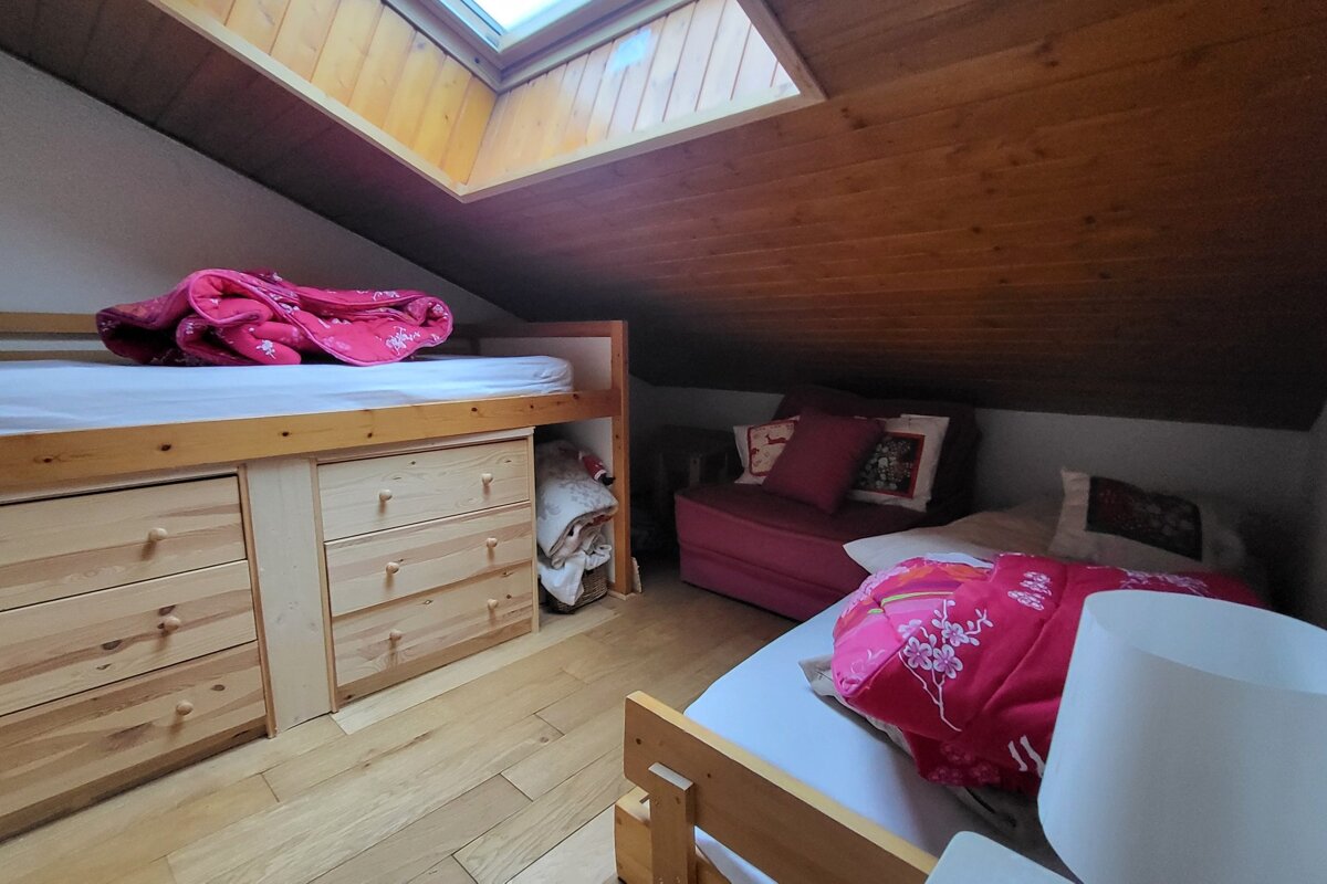 Cozy attic bedroom with a skylight, wooden ceiling, high bunk bed with drawers, a sofa bed, and another bed, all with pink accents.