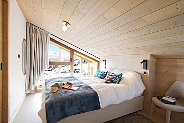 A cozy, light wood-paneled bedroom features a bed with blue accents and a large window framing a snowy mountain ski resort.
