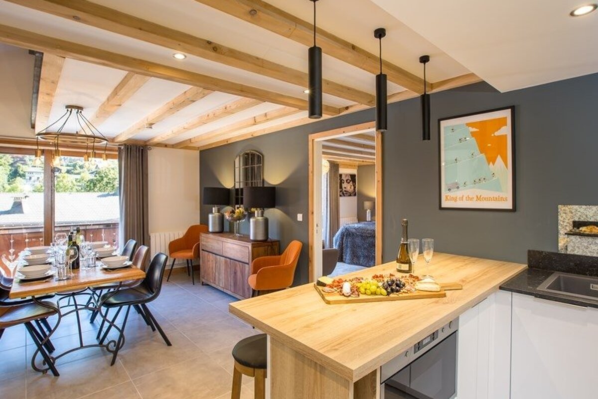 Modern open-plan kitchen and dining area with wooden ceiling beams, a set dining table, and a charcuterie board on the island counter.