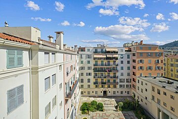 A vibrant cityscape of multi-colored residential buildings with shutters, surrounding a courtyard under a bright blue, cloudy sky.
