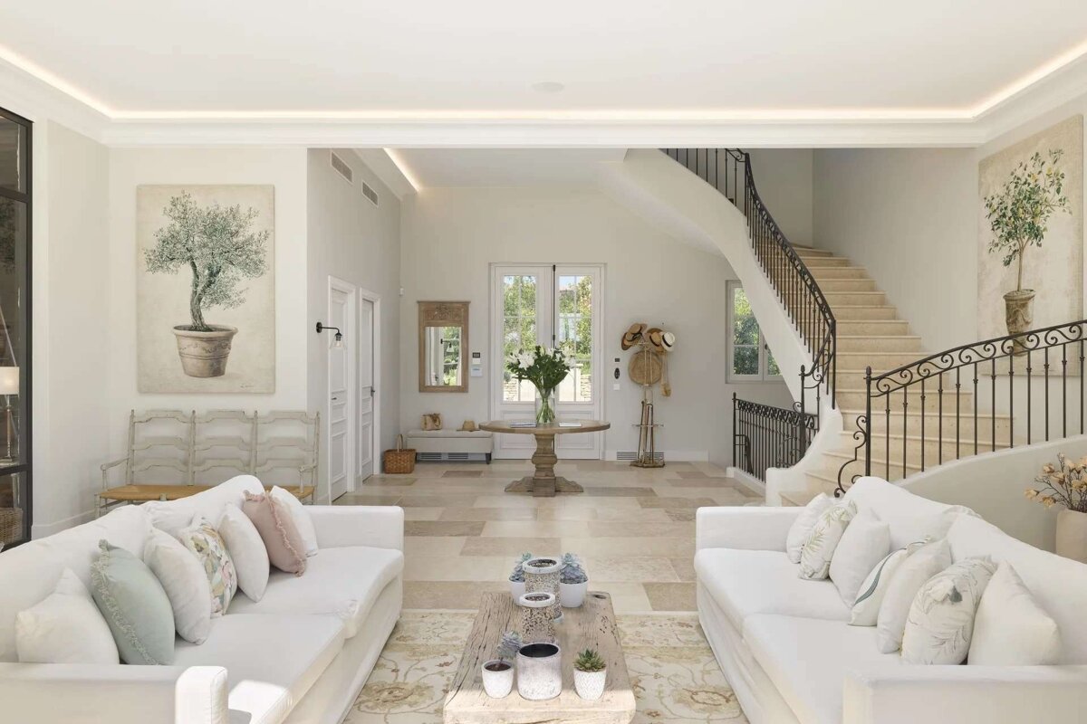 Elegant, spacious living room with white sofas, a grand curved staircase, light tiled floors, and botanical artwork.