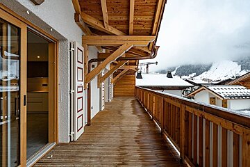 A long wooden balcony of an alpine chalet overlooking snowy roofs and misty mountains.