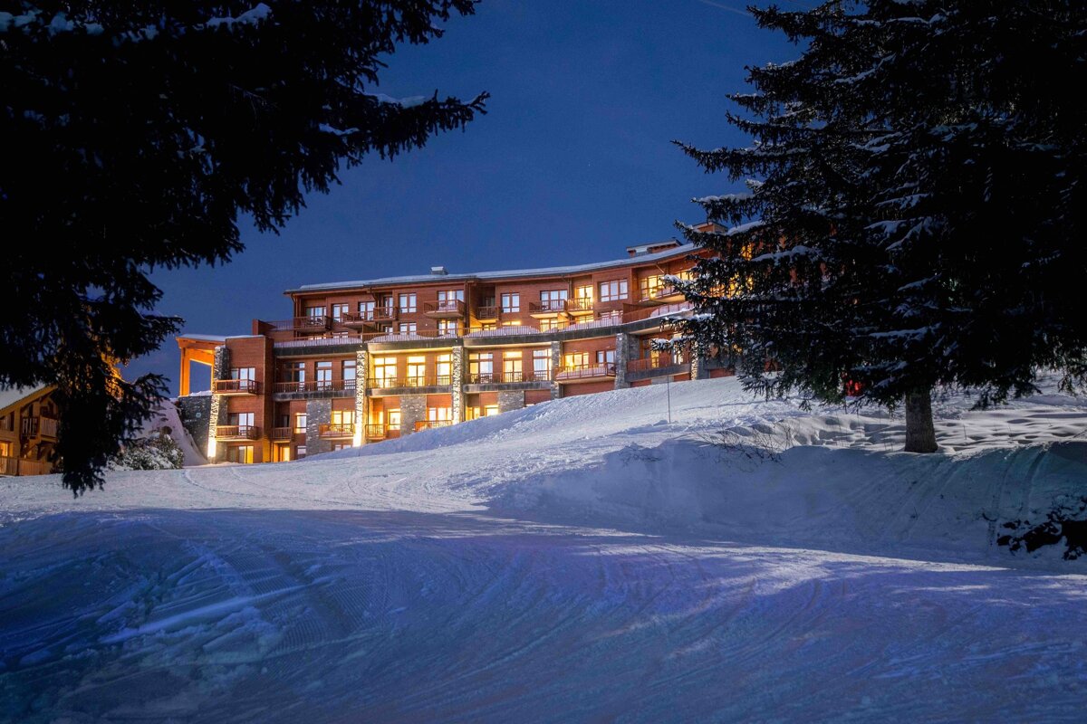 Brightly lit ski resort hotel nestled on a snow-covered slope at night, framed by dark evergreen trees under a deep blue sky.