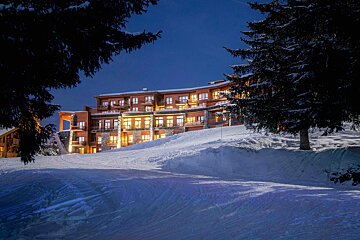 Brightly lit ski resort hotel nestled on a snow-covered slope at night, framed by dark evergreen trees under a deep blue sky.