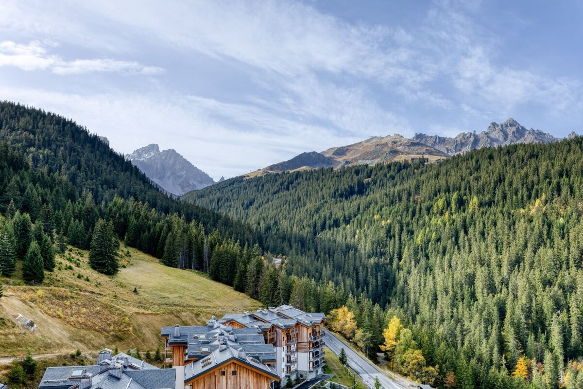 Panoramic view of a mountain valley with dense pine forests, rugged peaks, and a cluster of wooden chalets below a cloudy sky.