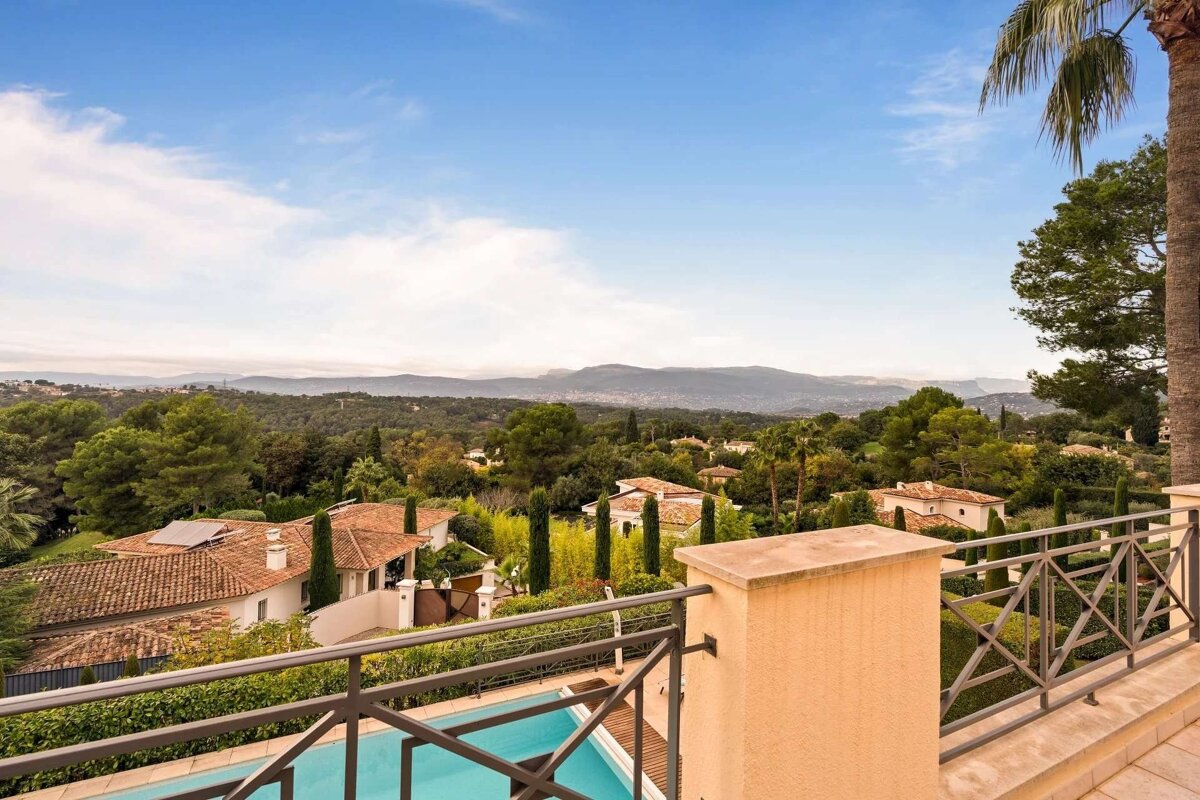 A scenic view from a balcony overlooking a pool, houses with tiled roofs, lush green trees, and distant mountains under a blue sky.