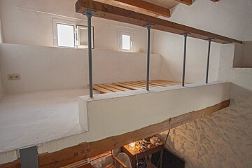 A bright, minimalist mezzanine level features a wooden bed frame, white walls, and small windows, supported by beams and metal poles, overlooking a lower room.
