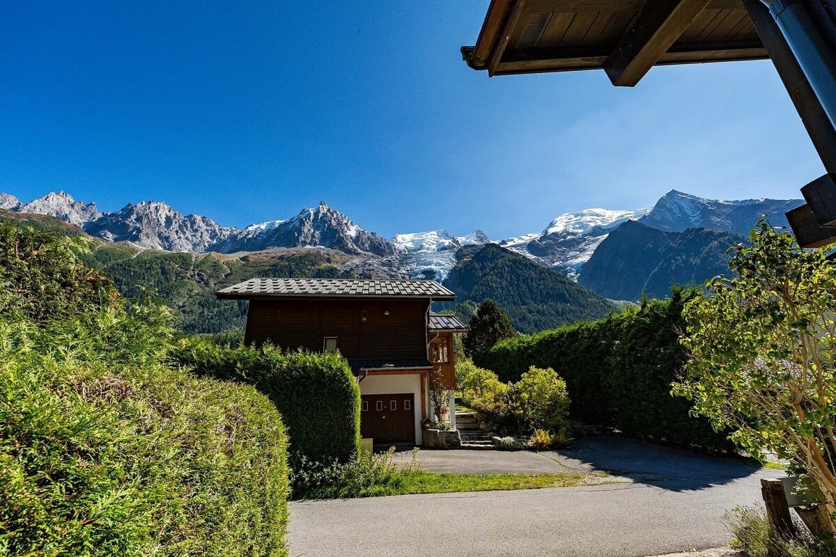 A wooden chalet with a driveway, framed by green hedges, sits before majestic snow-capped mountains under a brilliant blue sky.