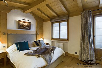 A rustic chalet bedroom featuring warm wood paneling, a large bed with a fur throw and teal pillows, and bright windows.