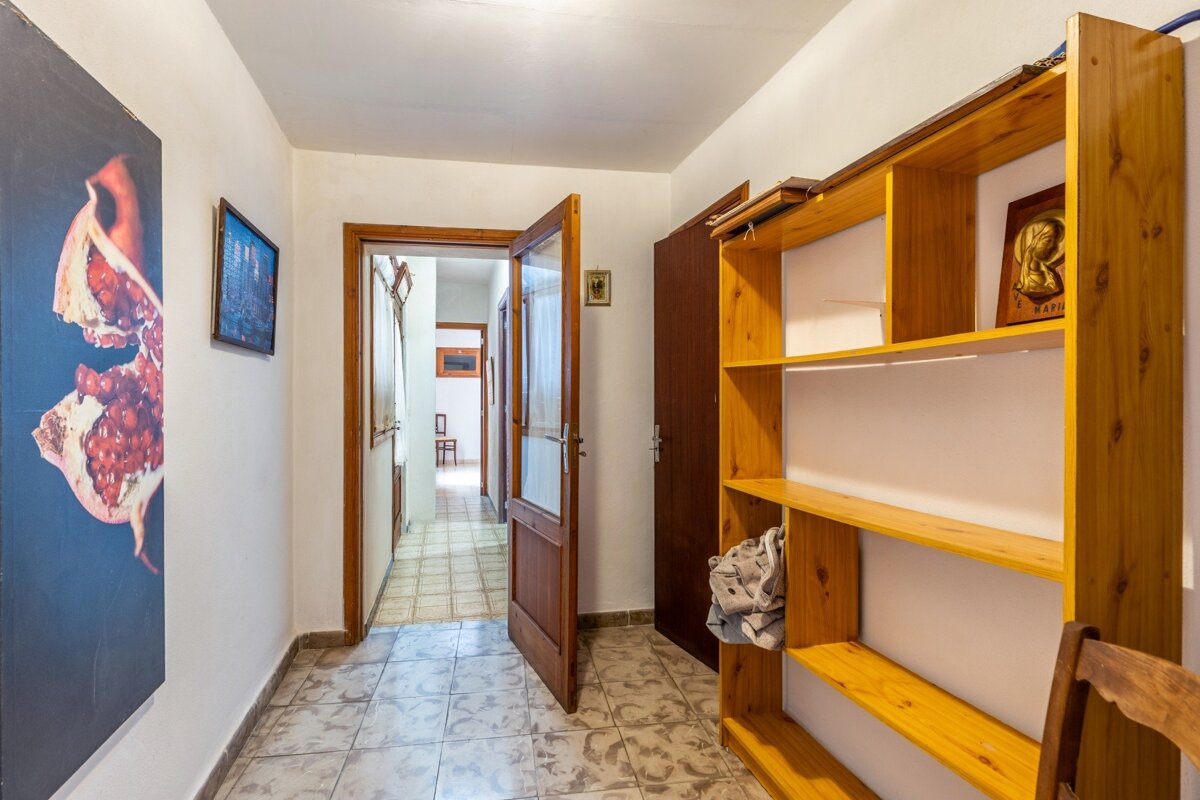 A tiled hallway with open wooden doors, a large pomegranate artwork on the left, and an empty wooden bookshelf on the right.