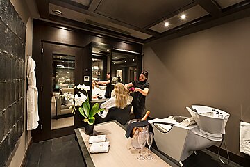 A stylist dries a client's hair in a luxurious, modern salon. Dark walls, orchids, champagne, and robes suggest a high-end spa experience.
