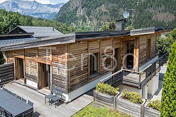 Modern wooden chalet with a large deck and balcony, set against a backdrop of green mountains.