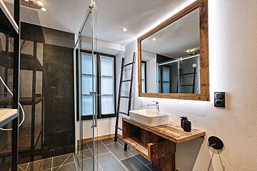 A chic bathroom features a glass shower, dark tiled walls, a rustic wooden vanity with a vessel sink, and a large illuminated mirror.