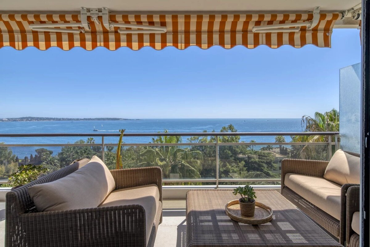 Balcony with rattan seating, coffee table, and a striped awning overlooking a vast blue ocean with distant boats under a clear sky.