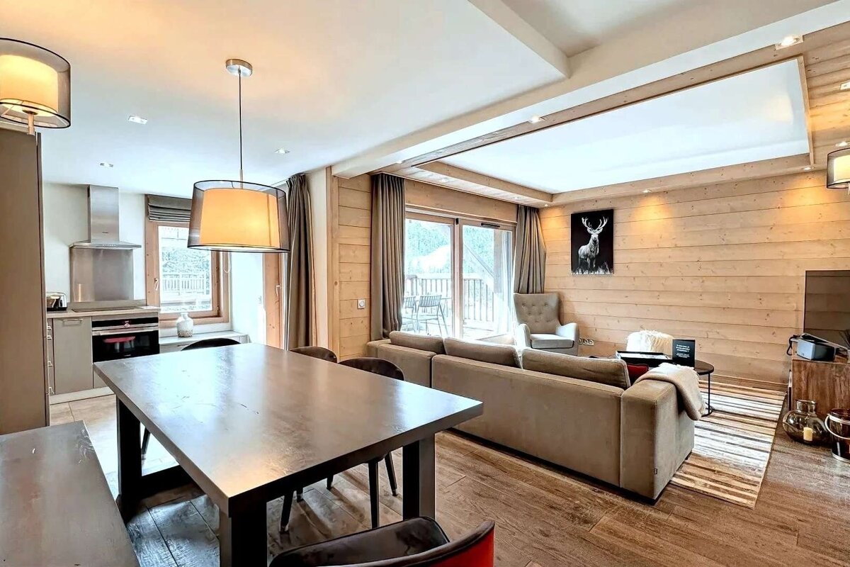 A modern open-plan living/dining/kitchen in a wood-paneled chalet with balcony access, featuring contemporary furnishings and deer art.