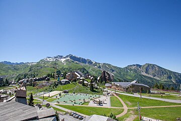 Scenic mountain resort village with green slopes, distant snow-capped peaks, buildings, a playground, and clear blue skies.