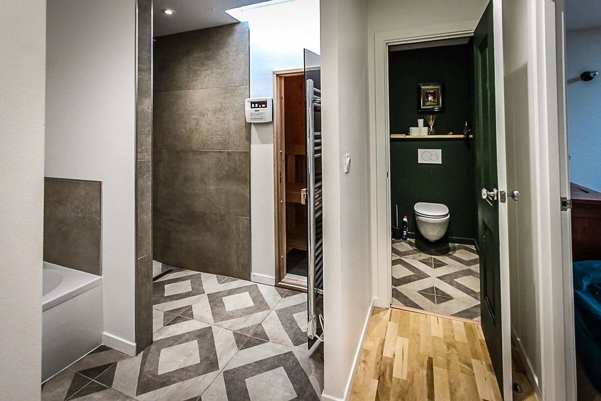 A modern bathroom/hallway area featuring geometric tiled floors, a sauna, a bathtub, and a dark green toilet room with wood flooring.