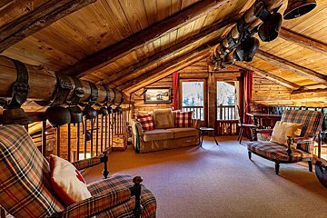 Cozy rustic wood-paneled chalet attic with plaid furniture, antique cowbells hanging from beams, and large windows showing snowy views.