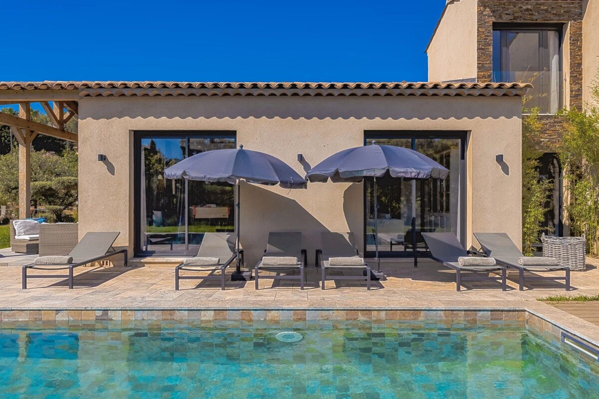 A bright swimming pool with gray lounge chairs and umbrellas lined up in front of a modern villa.