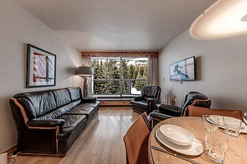 A cozy living/dining room features black leather furniture, a dining table, a TV, and a large window revealing a snowy mountain forest view.