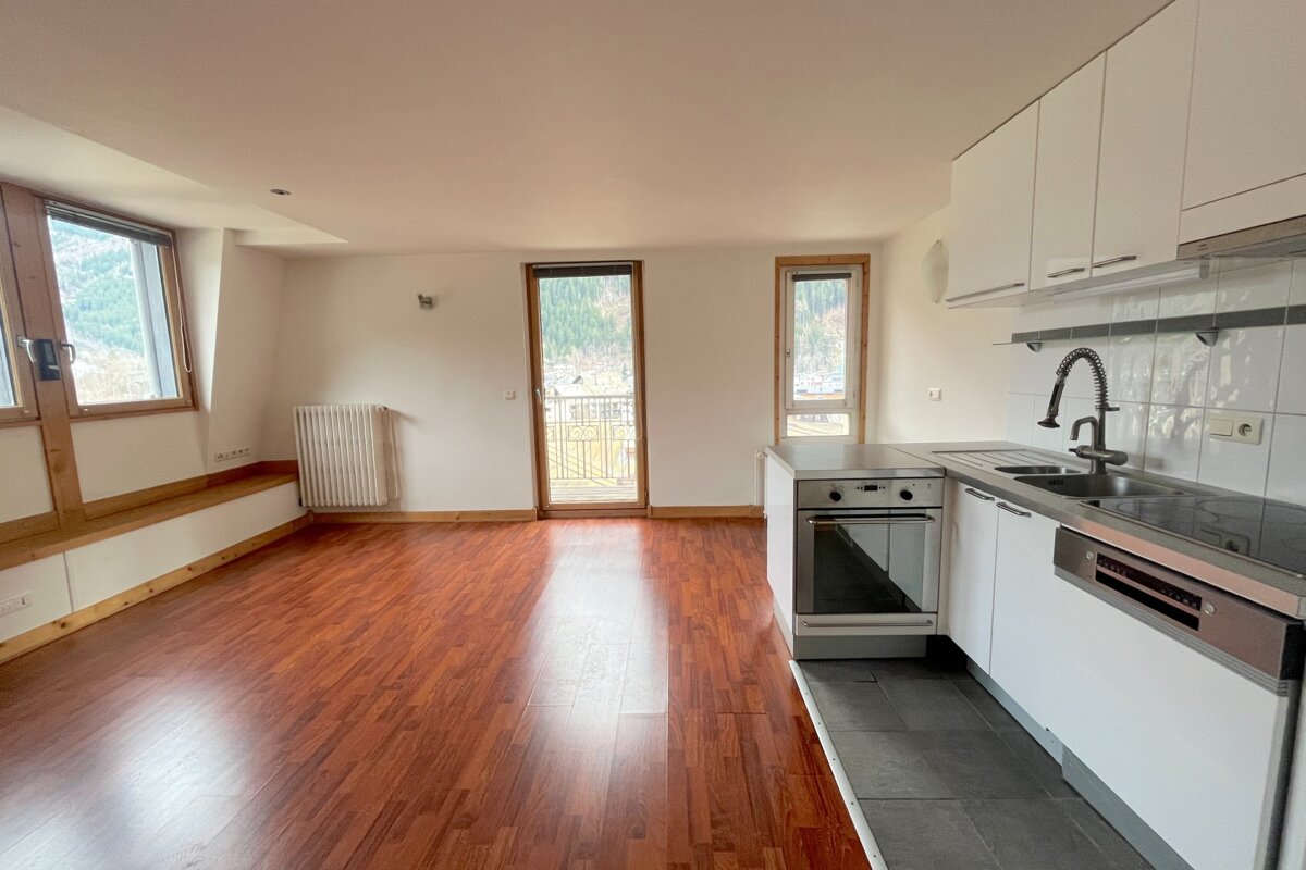 Bright, empty studio apartment with wood floors, a modern kitchen, built-in bench, and windows/balcony offering mountain views.