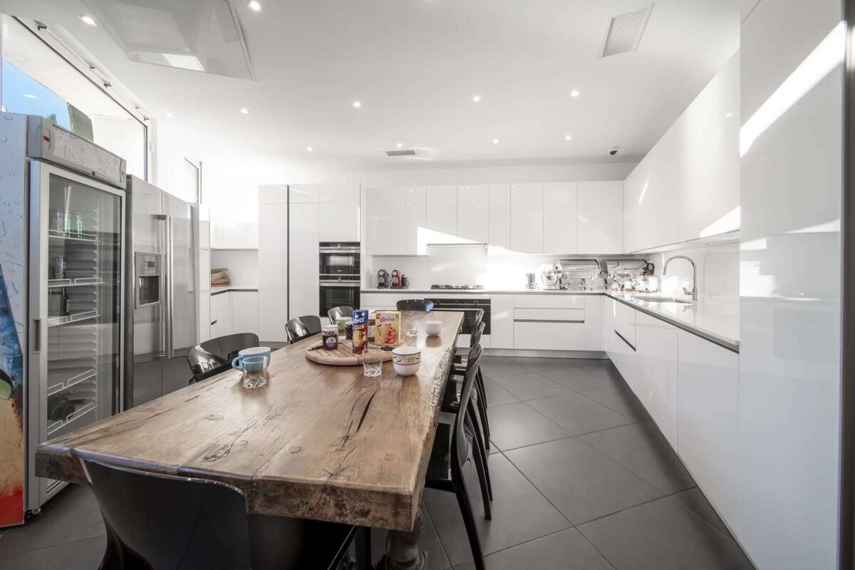Bright, modern kitchen with glossy white cabinets, dark tiled floor, and a large rustic wooden dining table set with breakfast items.