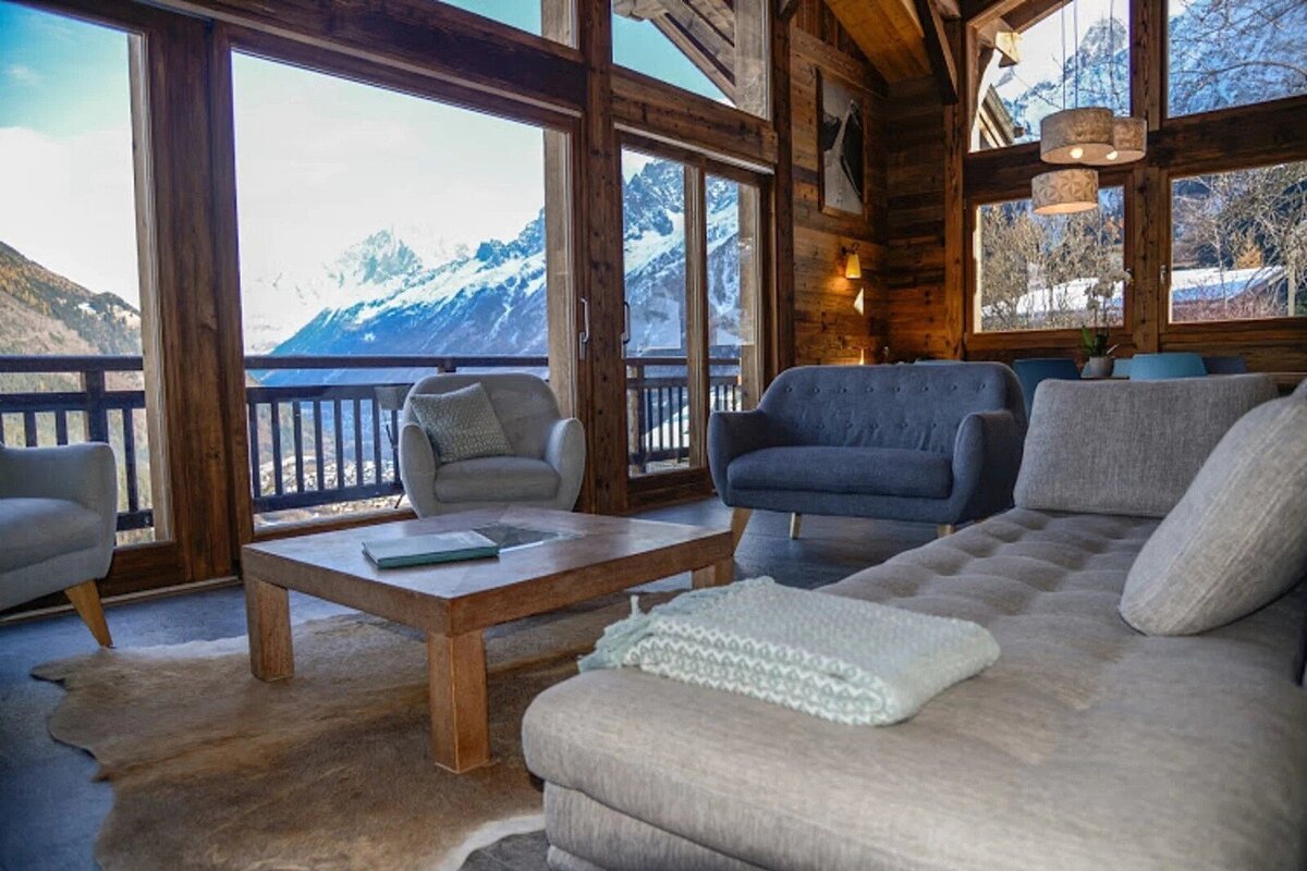 Cozy, rustic living room with stunning floor-to-ceiling views of snowy mountains. Features wooden beams, comfortable seating, and a coffee table.
