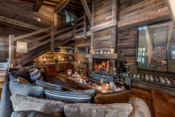 Cozy wooden chalet living room with a glowing fireplace, a large plush sectional sofa, and rustic stairs leading to a loft.