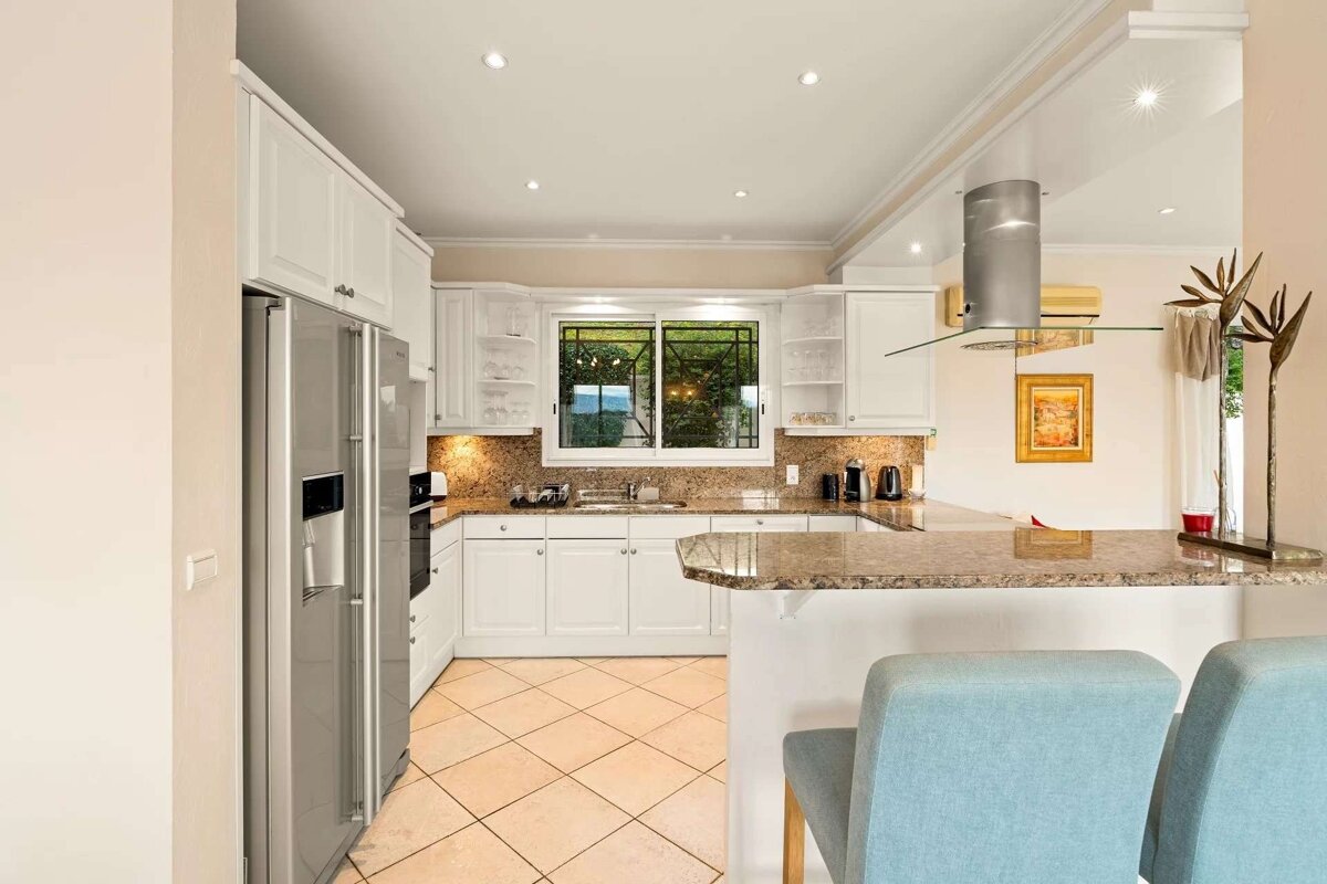 A bright, modern kitchen features white cabinetry, granite countertops, stainless steel appliances, tiled floors, and a breakfast bar with blue stools.