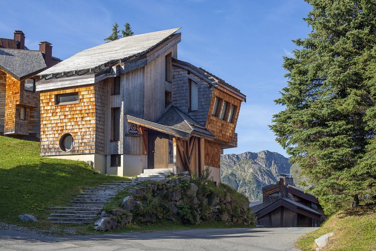 A uniquely designed wooden house with shingle siding and angled roofs sits on a grassy slope with mountains in the background under a blue sky.
