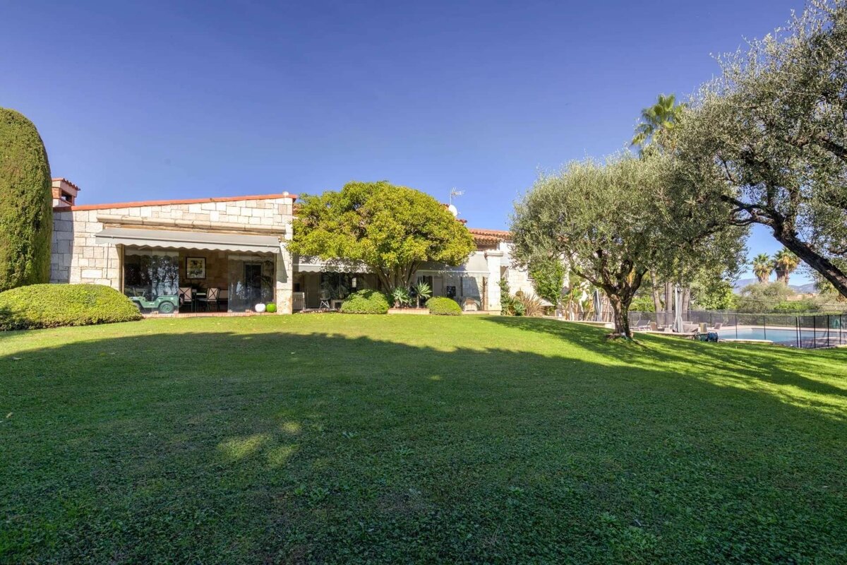 Sunny view of a stone house with a large green lawn, scattered trees, and a swimming pool under a clear blue sky.