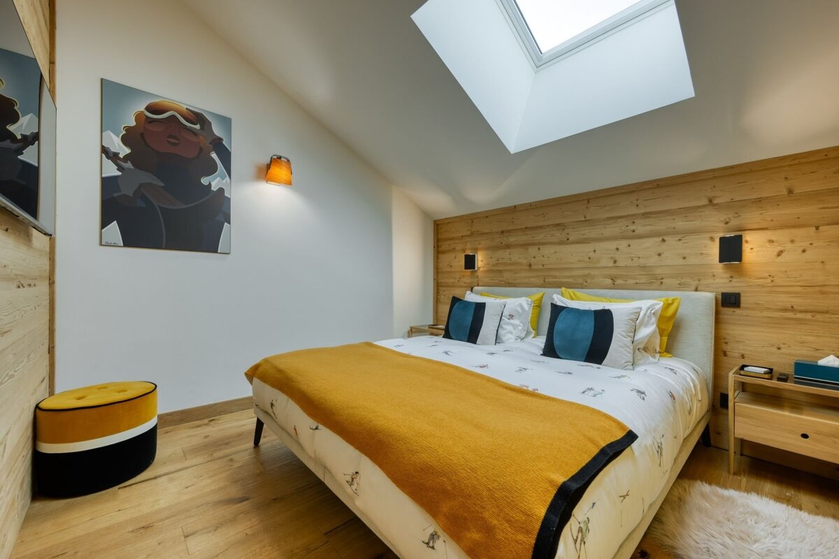 A modern, cozy bedroom with a bed featuring a yellow throw and blue pillows. It has a wooden accent wall, a skylight, and ski-themed artwork.
