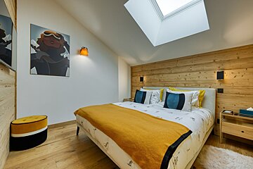 A modern, cozy bedroom with a bed featuring a yellow throw and blue pillows. It has a wooden accent wall, a skylight, and ski-themed artwork.