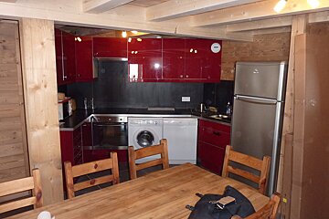 A cozy kitchen with bright red cabinets, black countertops, and integrated appliances like a washer/dryer, oven, and fridge. A wooden dining table fills the foreground.