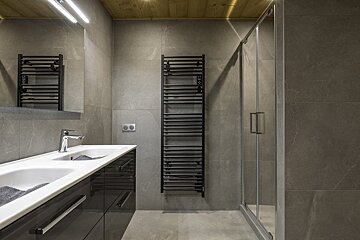 Modern gray-tiled bathroom with a double vanity, black towel warmer, glass shower, and warm wooden ceiling accents.
