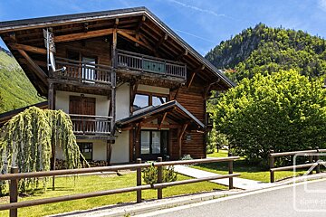 A large, rustic wooden chalet with balconies, nestled in a green mountain landscape. A fence, lawn, and trees are in the foreground under a blue sky.