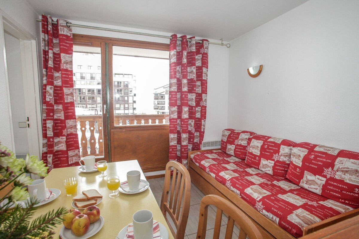 A cozy ski-themed apartment with breakfast on the dining table, a red patterned sofa and curtains, plus a balcony overlooking buildings.