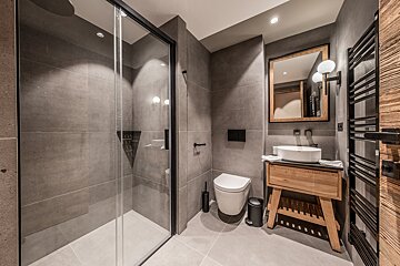 A sleek, modern bathroom with grey tiled walls, a glass shower, wooden vanity, white sink, and black fixtures, creating a minimalist and warm feel.
