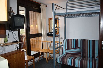 A compact room features a metal bunk bed, a striped futon, a wall-mounted TV, and wooden chairs, with a balcony door providing light.