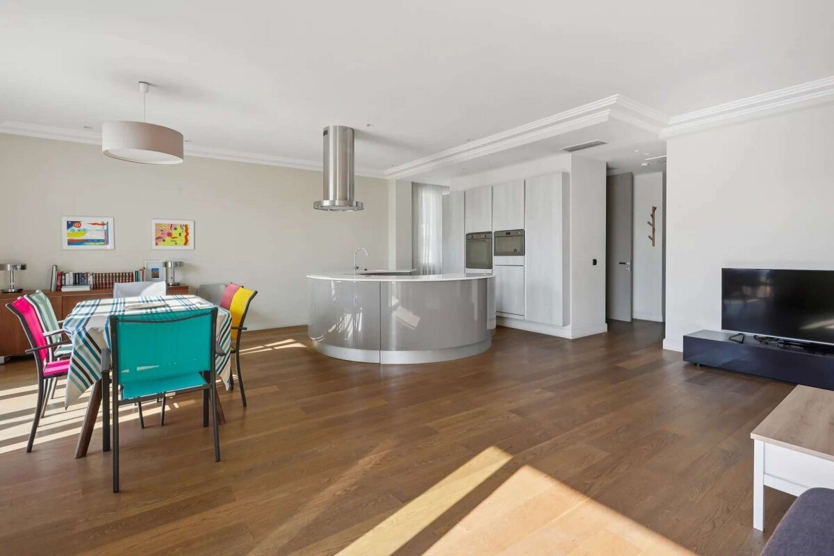 An open-plan living area with a colorful dining table, a modern kitchen featuring a curved island, and a television, all on a wooden floor.