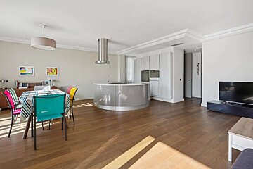 An open-plan living area with a colorful dining table, a modern kitchen featuring a curved island, and a television, all on a wooden floor.
