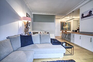 A stylish open-plan living room with a light gray sectional sofa, merging into a modern kitchen with a bar counter and stools. Bright and contemporary.