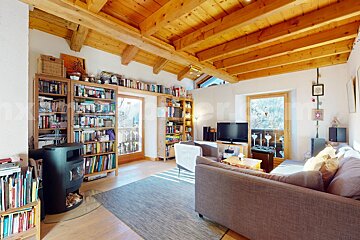 Cozy living room with a wooden beamed ceiling, extensive bookshelves, a wood stove, and comfy seating, opening to balconies with mountain views.