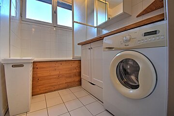 A clean bathroom featuring a front-load washing machine, white cabinets, a laundry hamper, tiled floor, and a bathtub with wooden paneling.
