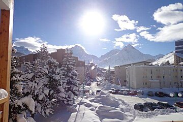A bright, sunny winter day in a snow-covered mountain village. Buildings, snow-laden pine trees, and parked cars are visible, with majestic peaks in the background.