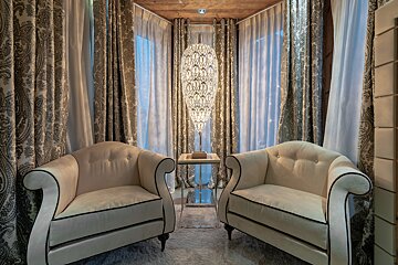 Two elegant beige armchairs face each other, separated by a tall ornate lamp, set against paisley curtains in a cozy nook.