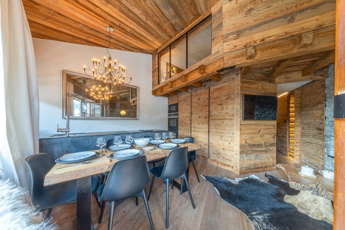 A cozy wooden chalet dining room featuring a rustic table with black chairs, a branching chandelier, and wood-paneled walls and ceilings.