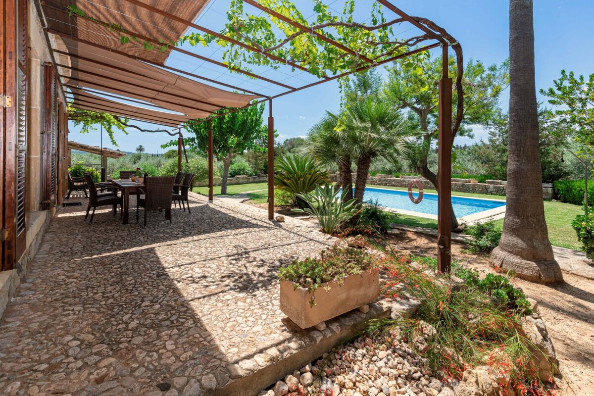 A sunny outdoor patio with a shaded dining area, cobblestone floor, lush garden, palm trees, and a sparkling blue swimming pool in the background.